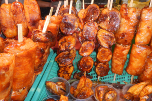 Ein Marktstand mit Spießen voller Hühnerflügel, verschiedenen Lebensmitteln in Plastikbehältern, mit Menschen und Flaschen im Hintergrund.