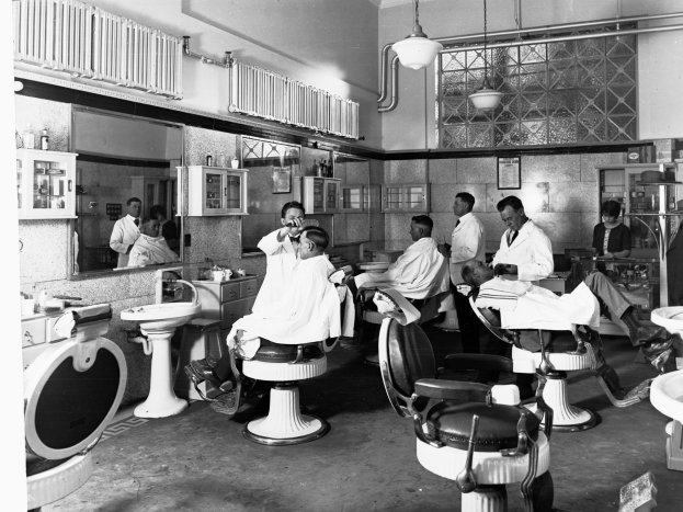 Schwarze und weiße Fotografie eines Friseursalons mit Menschen auf Stühlen und Stehenden, mit Waschbecken, Spiegeln, Schränken mit Flaschen und Deckenleuchten.