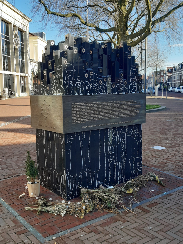 Denkmal in einem Stadtplatz für Holocaust-Opfer, mit einem Baum davor, einer Topfpflanze, verstreuten trockenen Blättern und Gebäuden, Fahrzeugen, Pfählen und Bäumen im Hintergrund.