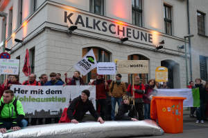 Eine Gruppe von Menschen steht vor einem Gebäude und hält Schilder und Plakate in die Höhe, während zwei Personen im Vordergrund sitzen und ein Müllcontainer rechts daneben steht, während sie bei einer Demonstration in Deutschland protestieren, wobei Gebäude, Fenster, Lichter und Schilder im Hintergrund zu sehen sind.