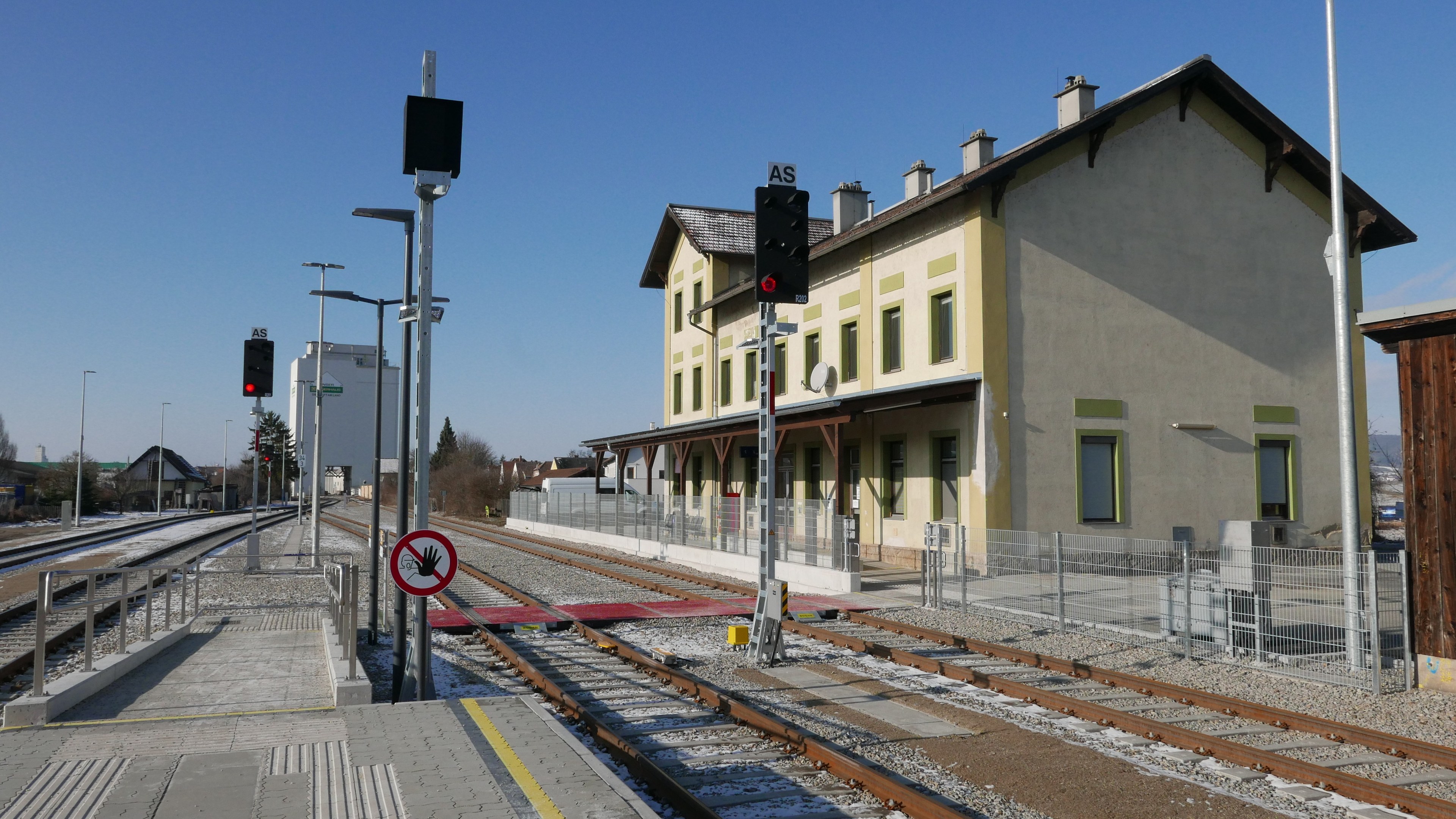 Ein Bahnhof mit einem Zug auf den Schienen, umgeben von Gebäuden, Straßenlaternen, Straßenlaternen, Verkehrspfosten, Verkehrssignalen, Schildern, Zäunen, Bäumen und einem klaren blauen Himmel.