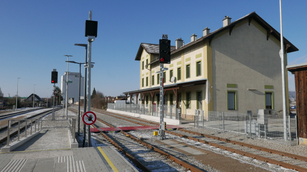Ein Bahnhof mit einem Zug auf den Schienen, umgeben von Gebäuden, Straßenlaternen, Straßenlaternen, Verkehrspfosten, Verkehrssignalen, Schildern, Zäunen, Bäumen und einem klaren blauen Himmel.