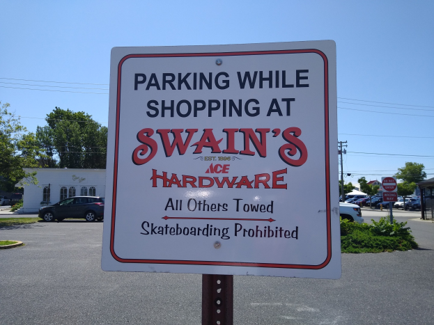 Parkplatz mit einem Schild, das 'Parken während des Einkaufs bei Swain's Hardware' anzeigt, im Vordergrund, Fahrzeuge auf der Straße, Pflanzen, Bäume, Gebäude, Pfosten, Drähte und einen klaren blauen Himmel im Hintergrund.