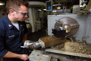 Ein Monteur mit Brille bedient eine Maschine in einer Fabrik, im Hintergrund sind Lampen, Rohre und andere Maschinen sowie ein Bildschirm zu sehen.