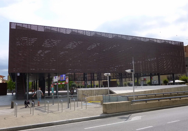 Großes modernes Gebäude mit zahlreichen Fenstern als neuer Eingang zur Stadt, flankiert von Straßeninfrastruktur, Fußgängern, Fahrzeugen und Bäumen unter einem bewölkten Himmel.