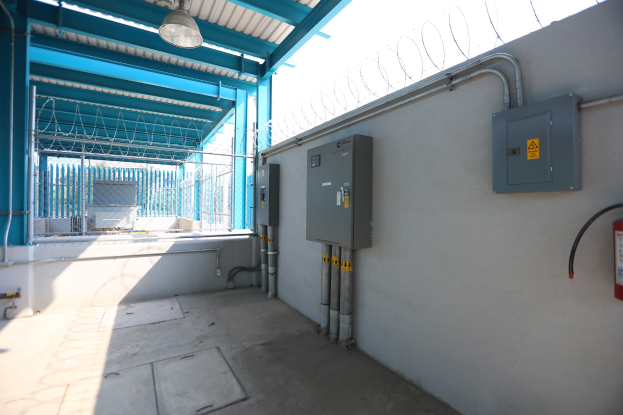 Ein Raum in einer elektrischen Umspannstation mit einem Feueröscher auf der rechten Seite, elektrischen Schaltkästen und Rohren an der Wand, einer Decke mit einer Lampe öber und einem Zaun im Hintergrund.
