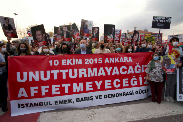 Eine Gruppe von Menschen mit Masken und Schildern und Transparenten protestiert vor einem Gebäude in Ankara, Türkei, gegen den Tod des türkischen Präsidenten Recep Tayyip Erdogan, mit Bäumen und Laternenmasten im Hintergrund unter einem klaren Himmel.