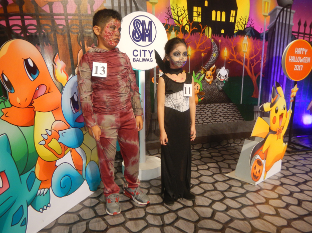 Zwei Kinder in Halloween-Kostümen stehen nebeneinander vor einer Wand mit einem Gemälde eines Gebäudes, Bäume, Laternenmasten und einem Zaun sowie Schildern mit Text und Tierbildern und festlicher Beleuchtung.