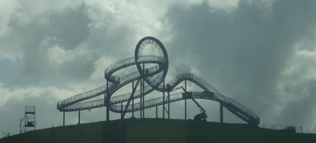 Eine Achterbahn auf dem Gipfel eines Hügels unter einem bewölkten Himmel, umgeben von Stützpfählen und anderen Strukturen.