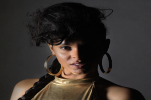 Eine Frau in einem goldenen Kleid und großen Creolen steht selbstbewusst vor einer Wand, ihre glatten Bob-Frisur ist ordentlich frisiert, trägt dezentes Make-up mit rosigem Rouge und glänzenden Lippen, schaut direkt in die Kamera mit einem entschlossenen Gesichtsausdruck.