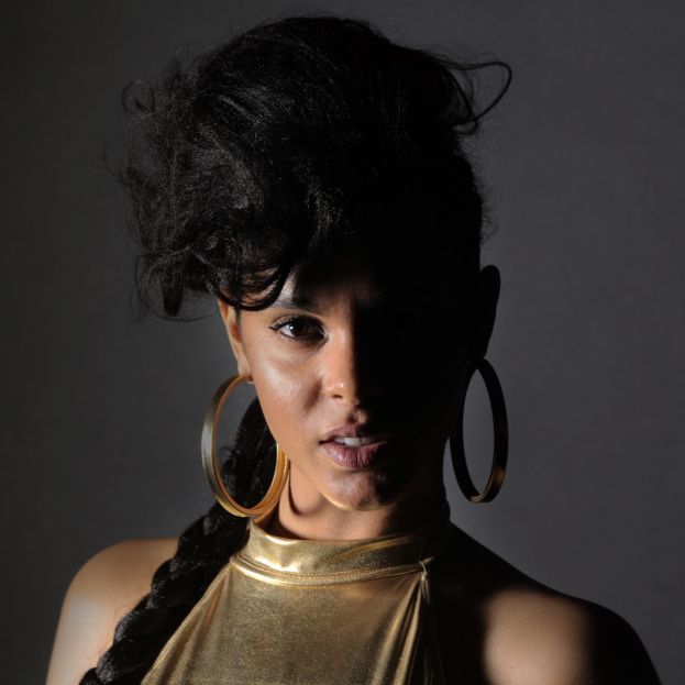 Eine Frau in einem goldenen Kleid und großen Creolen steht selbstbewusst vor einer Wand, ihre glatten Bob-Frisur ist ordentlich frisiert, trägt dezentes Make-up mit rosigem Rouge und glänzenden Lippen, schaut direkt in die Kamera mit einem entschlossenen Gesichtsausdruck.