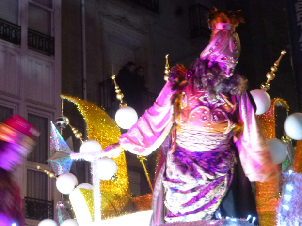 Ein Mann in einem Kostüm steht auf einem dekorierten, beleuchteten Festwagen in einem Stadtumzug, mit einem Gebäude im Hintergrund, das Fenster und Geländer hat.
