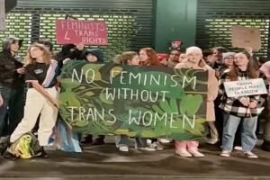 Eine Gruppe von Menschen hält ein Transparent mit der Aufschrift "Kein Feminismus ohne Trans-Frauen" und Schilder in einem öffentlichen Raum mit einer Wand und einem Rollladen im Hintergrund.