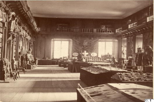 Ein altes Schwarz-Weiß-Foto eines Raumes voller Tische, Stühle, Skulpturen an den Wänden, Fenstern, Vorhängen, Geländern und anderen Gegenständen, mit dem Text "The Museum of Fine Arts, London" unten.