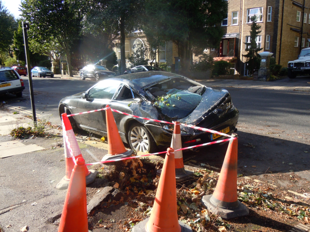 Ein besch├Ądigt auf der Stra├če stehendes Auto, mit orangefarbenen Verkehrskegeln abgesperrt, mit B├Ąumen, Geb├Ąuden und einem klaren blauen Himmel im Hintergrund.