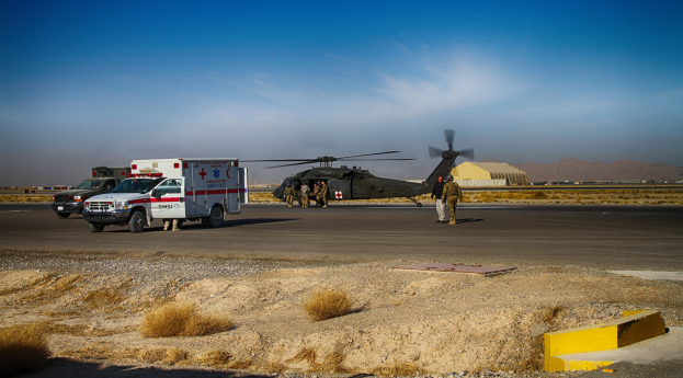 Hubschrauber auf gröner Wiese neben Krankenwagen mit Menschen in der Nähe, Hügel und klarer blauer Himmel im Hintergrund.