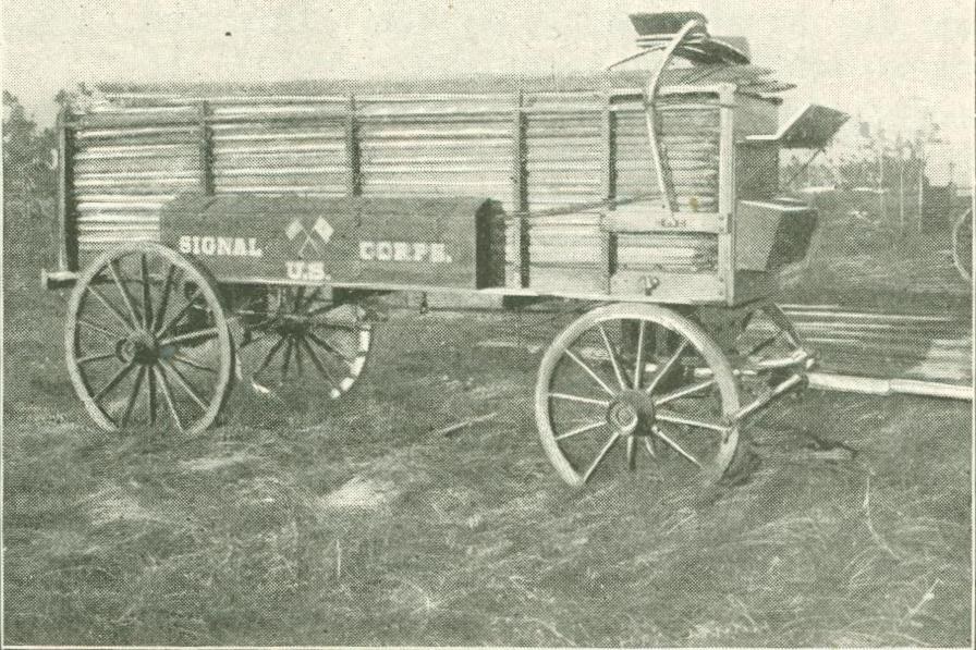 Schwarz-weiß-Foto eines Wagens mit einem Schild, umgeben von Gras und Bäumen unter einem klaren Himmel, beschriftet mit 'Signal U.S. Corp. Wagon' unten.