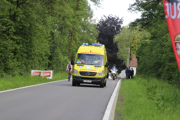 Ambulanz auf einer Straße mit Fahrradfahrern daneben, umgeben von Gras, Bäumen, Häusern, Strommasten und einem klaren blauen Himmel.