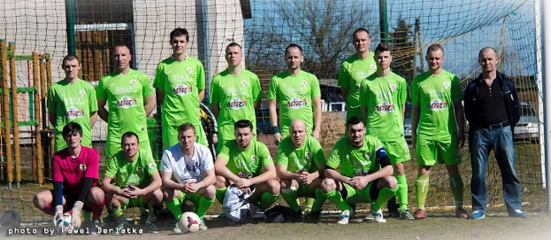 Eine Gruppe von Männern in grünen T-Shirts steht und hockt auf einem Fußballfeld mit einem Ball vor ihnen, einem Torpfosten rechts daneben und einem Hintergrund aus Bäumen, Gebäuden, Pfählen, Fahrzeugen und dem Himmel, mit Text unten links.