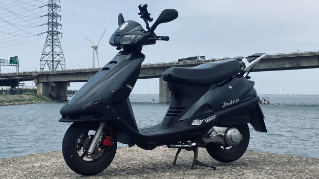 Schwarzer E-Scooter neben einem Gewässer mit einer Brücke, einem Turm mit Drähten, einer Windmühle und einem klaren blauen Himmel im Hintergrund geparkt.
