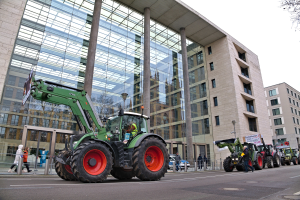 Traktorgruppe fährt auf einer Straße vor einem Gebäude, mit Fußgängern auf dem Gehweg und einem Baum auf der rechten Seite, während einer Demonstration.