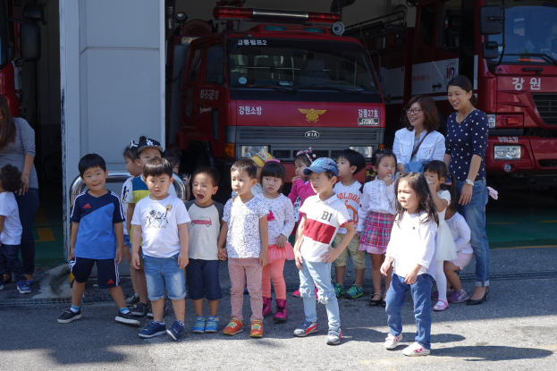 Eine Gruppe von Kindern vor einem Feuerwehrauto auf einer Feuerwache, einige tragen Mützen und andere Feuerwehrautos sind im Hintergrund zu sehen.