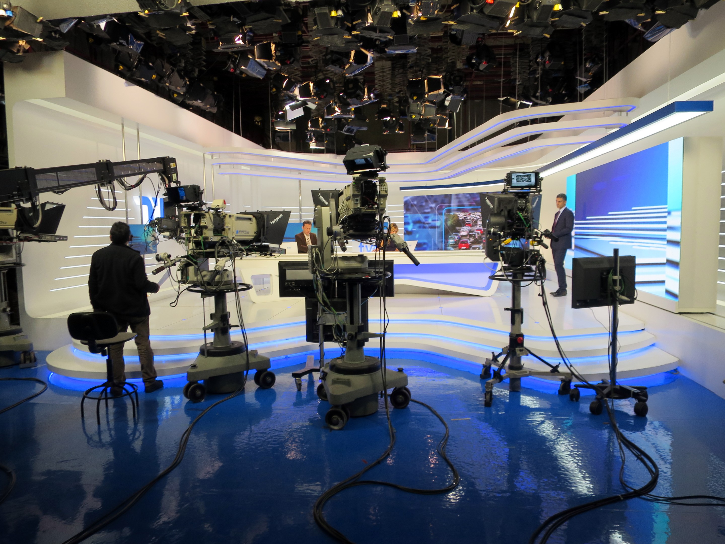 Ein TV-Studio mit mehreren Kameras auf dem Boden, einer sitzenden Person im Vordergrund und einigen stehenden Personen im Hintergrund, sowie an der Decke montierten Lichtern für eine Livesendung.