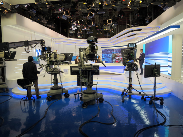 Ein TV-Studio mit mehreren Kameras auf dem Boden, einer sitzenden Person im Vordergrund und einigen stehenden Personen im Hintergrund, sowie an der Decke montierten Lichtern für eine Livesendung.