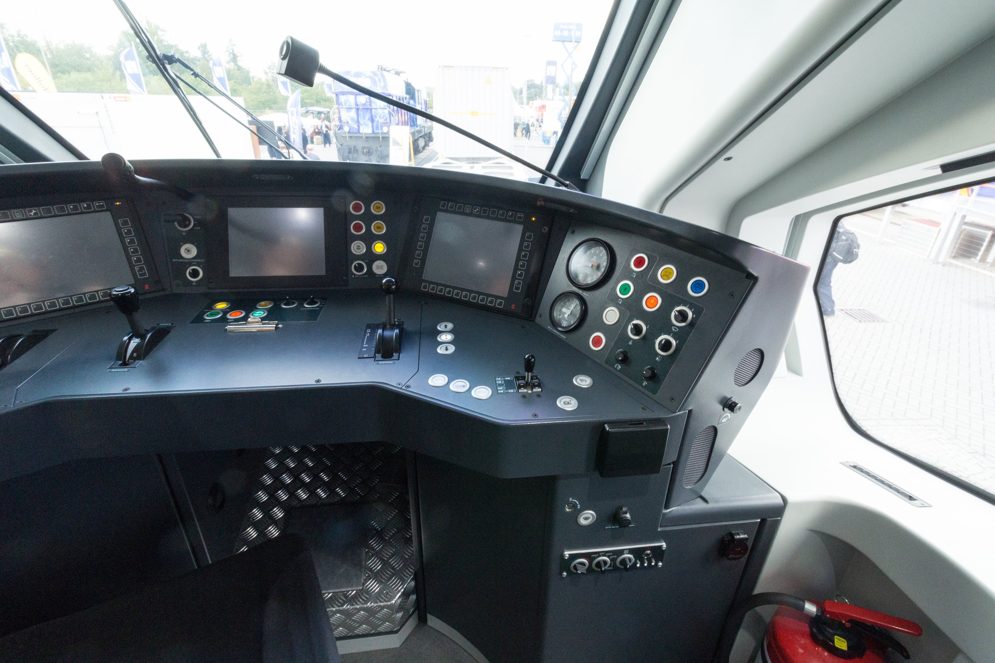 Cockpit eines Zuges mit Feuerlöscher im Vordergrund, das eine Glasfensteransicht von Menschen, Fahrzeugen, Bäumen und anderen Objekten draußen zeigt.
