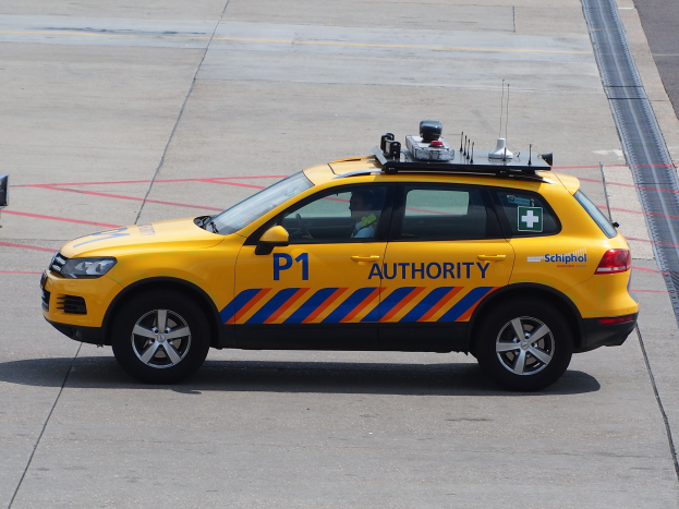 Gelbes Polizeiauto mit "Taxi"-Text an der Seite auf dem Rollfeld eines Flughafens geparkt, eine Person im Inneren und ein weiteres Fahrzeug links daneben.