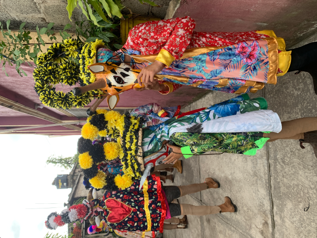 Eine Gruppe von Menschen in farbenfrohen Kostümen steht zusammen vor einem Gebäude, mit verschiedenen Gegenständen, Bäumen, Gebäuden und einem klaren blauen Himmel im Hintergrund während eines Karnevalsumzugs.