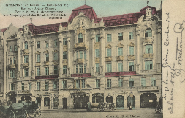 Eine alte Postkarte des Grand Hotel de Russie in Berlin, Deutschland, zeigt ein mehrstöckiges Gebäude mit zahlreichen Fenstern, eine Gruppe von Menschen am Boden, Pferdewagen, Straßenmasten und Schilder unter einem bewölkten Himmel.