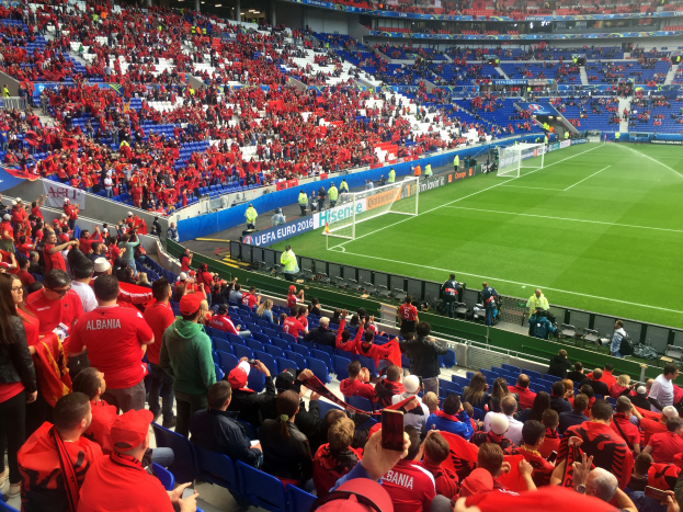 Ein volles Stadion mit Zuschauern, einige sitzen und einige stehen, mit Bannern, einem Torpfosten und Deckenleuchten, während die Menge ungeduldig auf den Beginn eines Fußballspiels wartet.
