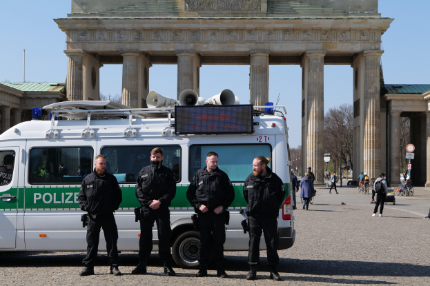Eine Gruppe von Polizisten in schwarzen Uniformen vor dem Brandenburger Tor in Berlin, mit einem weißen und grünen Fahrzeug im Vordergrund, Passanten, Radfahrer, Schilder, Bäume und das Brandenburger Tor im Hintergrund.