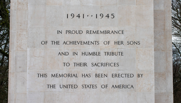 Ein Denkmal mit einer Tafel, auf der 'In stolzem Gedenken an die Taten ihrer Söhne und in Demut zur Ehrung ihres Opfers' steht, vor einem Hintergrund aus Bäumen und einem klaren blauen Himmel.