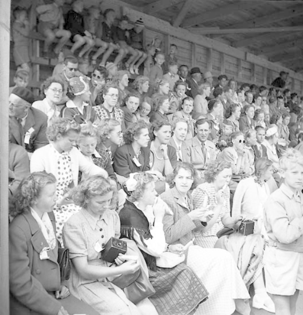 Schwarzes und weißes Foto einer Menschenmenge, die in einem Stadion mit einer hölzernen Wand und einem Dach sitzt, wobei einige Kameras halten.
