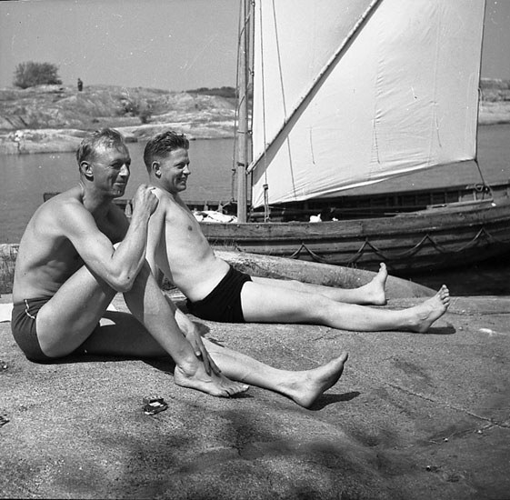 Zwei Männer in Badebekleidung sitzen auf einem Strand neben einem Segelboot, mit blauem Wasser, grünen Bäumen und einem weißen Himmel im Hintergrund.