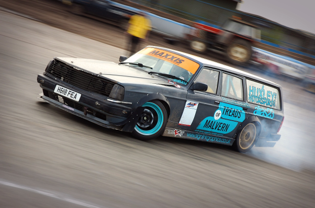 Ein Auto auf einer Rennstrecke, das Rauch ausstößt, umgeben von anderen Fahrzeugen und Zuschauern, was auf hohe Geschwindigkeit hinweist.