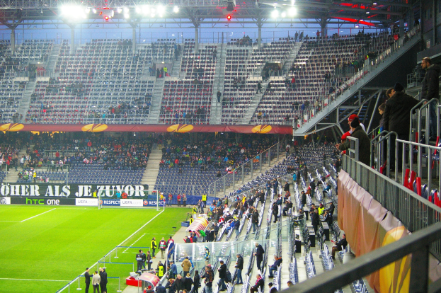 Großes Stadion voller Zuschauer bei einem Fußballspiel, mit sitzenden und stehenden Personen, einem Geländer, einer rechten Banner und einem leeren beleuchteten Stadion im Hintergrund.