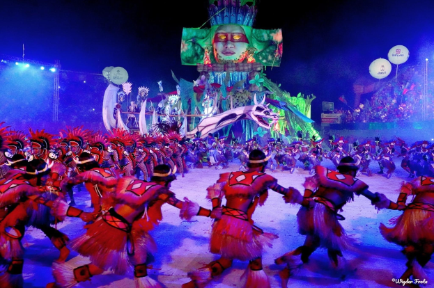 Eine Gruppe von Menschen in bunten Kostümen tanzt auf einer Bühne vor einem sitzenden Publikum, mit Lichtern, Stangen, Ballons und einem Bildschirm, der eine Person in der Hintergrund zeigt, unter einem sichtbaren Himmel, bei der Rio-Karneval 2014.