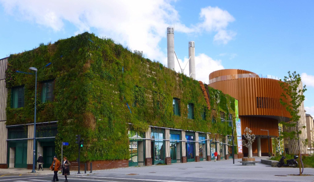 Großes nachhaltiges Gebäude mit Green Wall, umgeben von Straßeninfrastruktur, Fahrzeugen, Fußgängern, Bäumen und bewölktem Himmel.