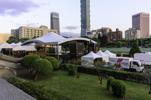 Gruppe von Zelten in einem Park umgeben von Grünanlagen, mit Menschen, Fahrzeugen, Gebäuden und einem klaren Himmel im Hintergrund, wahrscheinlich bei dem Hong Kong International Film Festival aufgenommen.