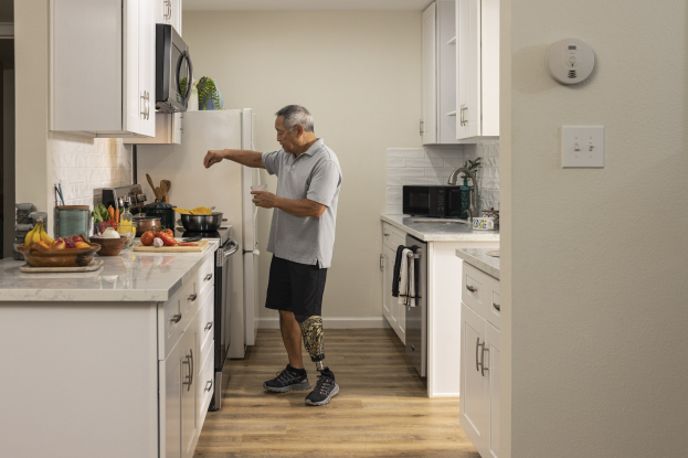 Ein Mann steht in einer Küche mit weißen Schränken, hält ein Glas in der Hand und ist von Obst, Gemüse, Schüsseln und Küchenutensilien auf einer Plattform zu seiner Linken umgeben, mit Schränken, Geräten und einer Vase im Hintergrund.