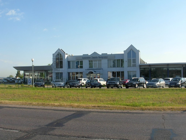 Autos vor einem Autohaus geparkt, mit Gebäuden, Straßenlaternen, Fahrzeugen, Schildern, Bäumen und bewölktem Himmel im Hintergrund.
