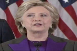 Hillary Clinton, gekleidet in ein lila Kleid und eine schwarze Jacke, spricht an einem Rednerpult mit Entschlossenheit und Stärke, neben einem Mann vor einer amerikanischen Flagge.