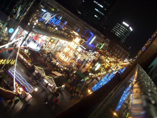 Eine belebte Stadtstraße bei Nacht voller Menschen, die unter den Lichtern der Gebäude und Stände gehen, mit einem dunklen Himmel im Hintergrund.