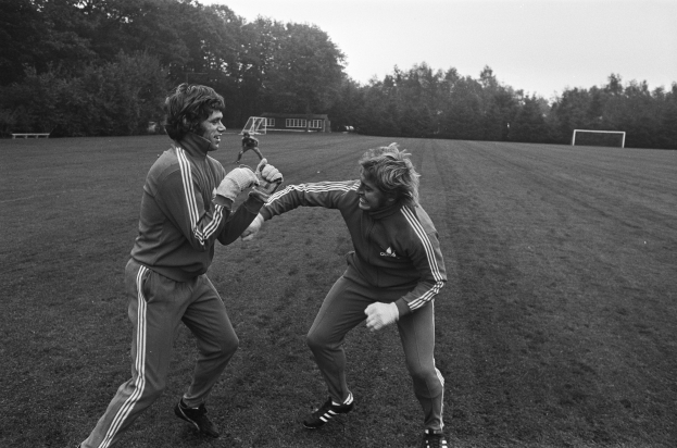 Zwei Männer in Schwarz-Weiß-Uniformen sind in eine körperliche Auseinandersetzung auf einem Fußballfeld verwickelt, umgeben von Bäumen und einem Schuppen im Hintergrund, beide tragen Handschuhe.