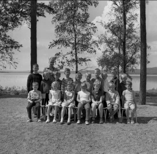 Schwarz-weiß-Fotografie von Kindern, die auf Stühlen sitzen und vor einem See stehen, mit Bäumen, Pflanzen, Hügeln und einem bewölkten Himmel im Hintergrund, beschriftet mit "1940er-Kinder am See."
