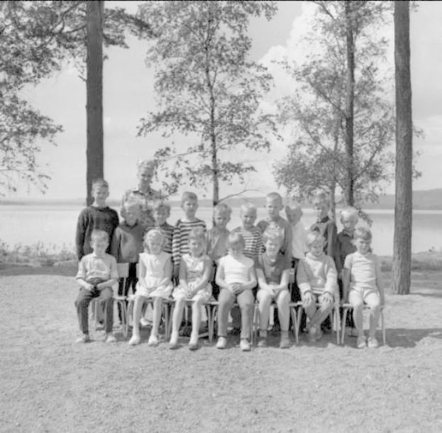 Schwarz-weiß-Fotografie von Kindern, die auf Stühlen sitzen und vor einem See stehen, mit Bäumen, Pflanzen, Hügeln und einem bewölkten Himmel im Hintergrund, beschriftet mit "1940er-Kinder am See."