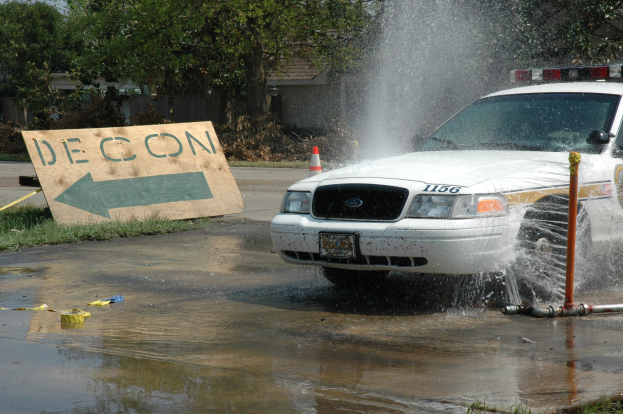 Ein Polizeiwagen fährt durch eine Pfütze, mit einem Schild, einem Verkehrskegel und anderen Gegenständen auf dem Boden, mit Bäumen, Gebäuden und einem klaren blauen Himmel im Hintergrund.